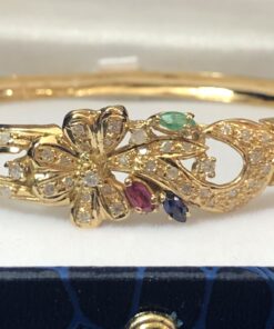Bonito brazalete rígido con motivos florales, enriquecido con 0'90 cts de brillantes, y detalles de rubí, esmeralda y zafiro naturales. Realizado en oro amarillo de ley 18 kts.